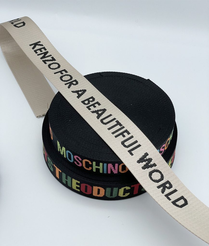 Beige custom jacquard webbing with black woven lettering crossing two rolls of black webbing